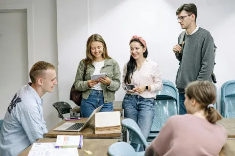 Grupo de jovens adultos a sorrir e conversar descontraidamente numa sala de aula, transmitindo leveza, boa disposição e espírito de partilha.