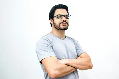 "Homem de braços cruzados com expressão confiante em frente a fundo branco
