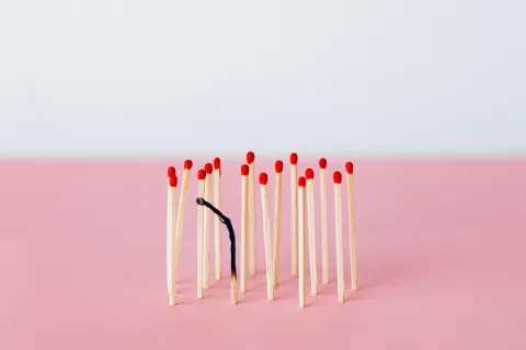 Um grupo de fósforos em pé sobre uma superfície rosa, com um fósforo queimado curvado no centro, simbolizando esgotamento ou burnout.