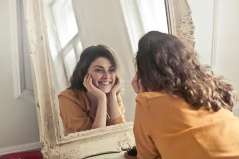 Mulher sorridente a olhar para si própria ao espelho com expressão de alegria e autoaceitação, simbolizando autoestima e bem-estar emocional.