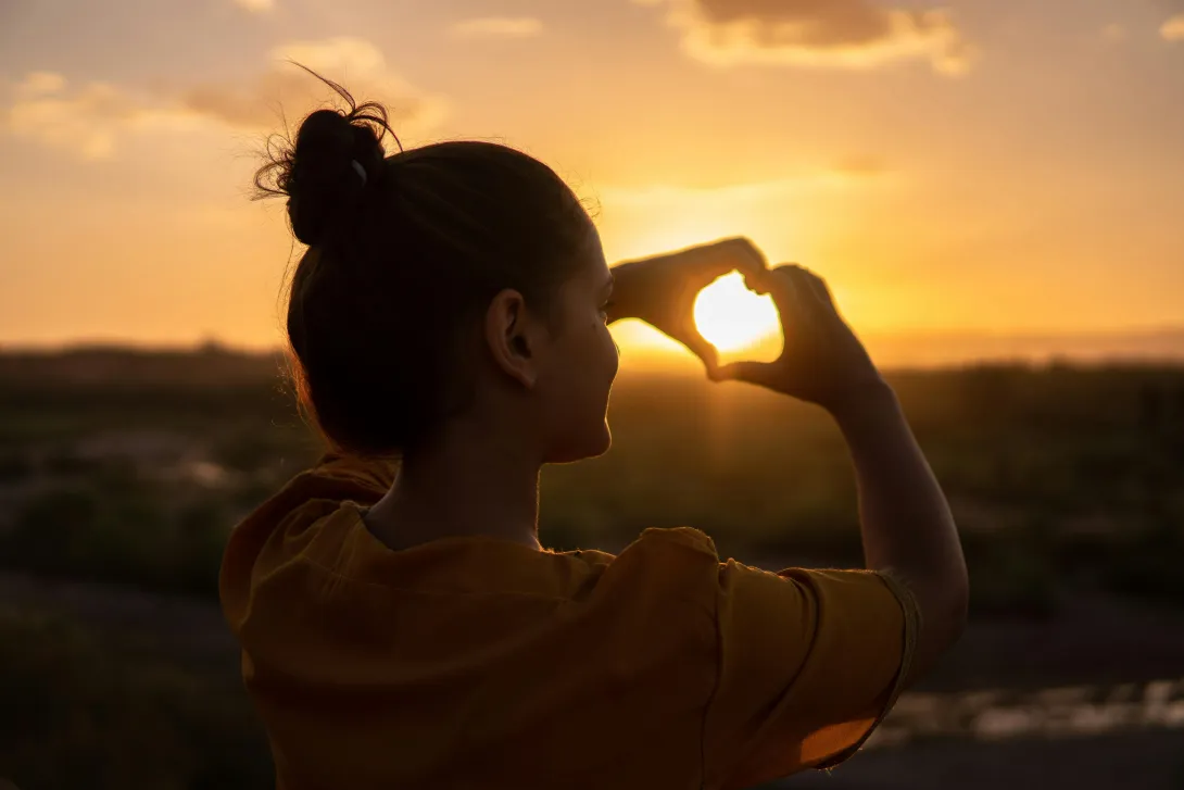 Pessoa a contemplar o pôr do sol e a formar um coração com as mãos, simbolizando amor‑próprio, gratidão e autoconhecimento.