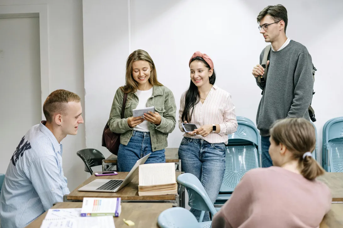 Grupo de jovens adultos a sorrir e conversar descontraidamente numa sala de aula, transmitindo leveza, boa disposição e espírito de partilha.