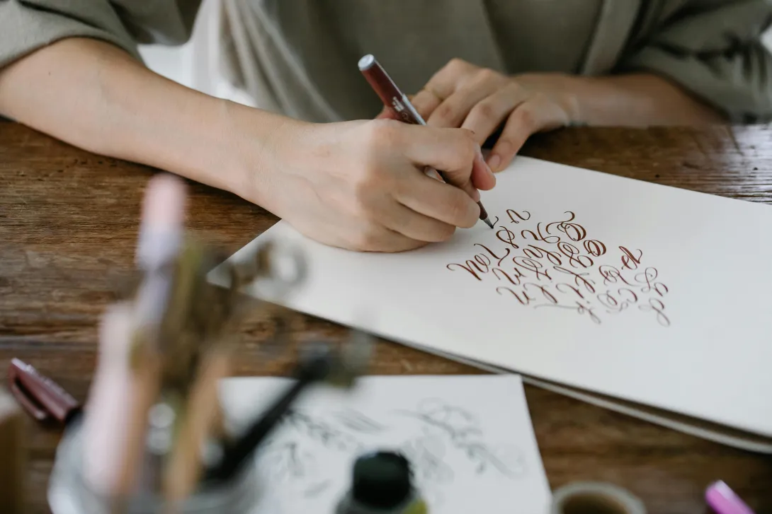 Pessoa a escrever com caneta de tinta castanha em folha branca, praticando caligrafia artística