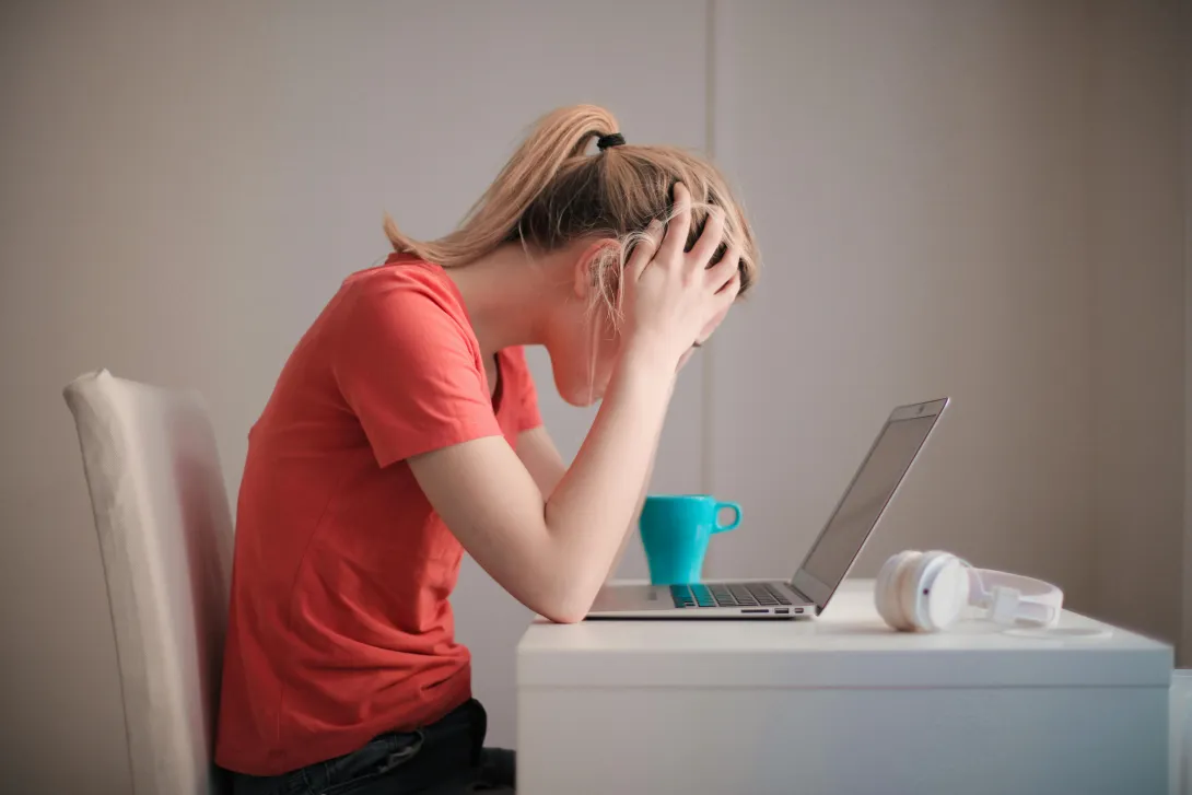 Mulher sentada à secretária com as mãos na cabeça em sinal de preocupação, olhando para o computador portátil, representando ansiedade e stress.